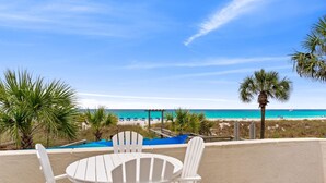 Une terrasse avec des chaises blanches et une table, d’où l’on peut admirer une plage bordée de palmiers et un ciel d’un bleu limpide.