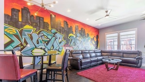 Un salon orné d’une fresque représentant un paysage urbain, avec un canapé en cuir noir, un tapis rouge et un coin repas comprenant des chaises et une table.