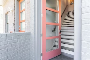 A red-framed glass door with a metal handle, a white brick wall, and a staircase with black steps. - City Heights- Rooftop Patio, Skyline Views, Sleeps 10 Near Broadway (Nashville)
