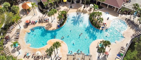 Eine Vogelperspektive zeigt ein Resort mit einem großen zentralen Pool, mehreren Liegebereichen und Palmen.