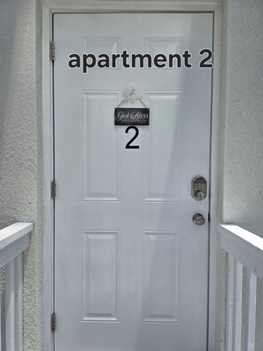 Eine weiße Tür mit einem Schild, auf dem 'Apartment 2' und 'Gott segne dieses Haus und alle, die es betreten' steht.