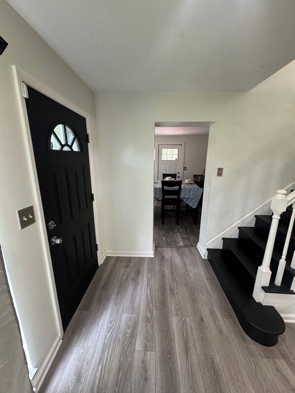 A hallway with a black door, a staircase, and a dining area in the background.