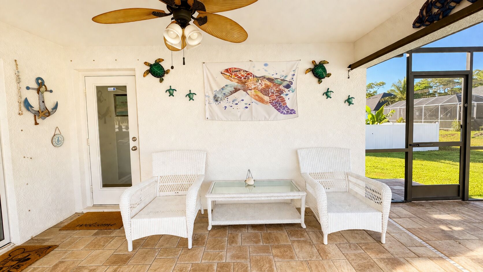 Une terrasse couverte avec deux fauteuils en osier blancs, une table à plateau en verre, un ventilateur de plafond et une banderole représentant une tortue de mer.