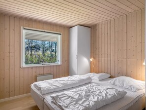 Ein Schlafzimmer mit Holzwänden, einem Fenster mit Jalousien, einem Bett mit weißer Bettwäsche und einer Nachttischlampe.