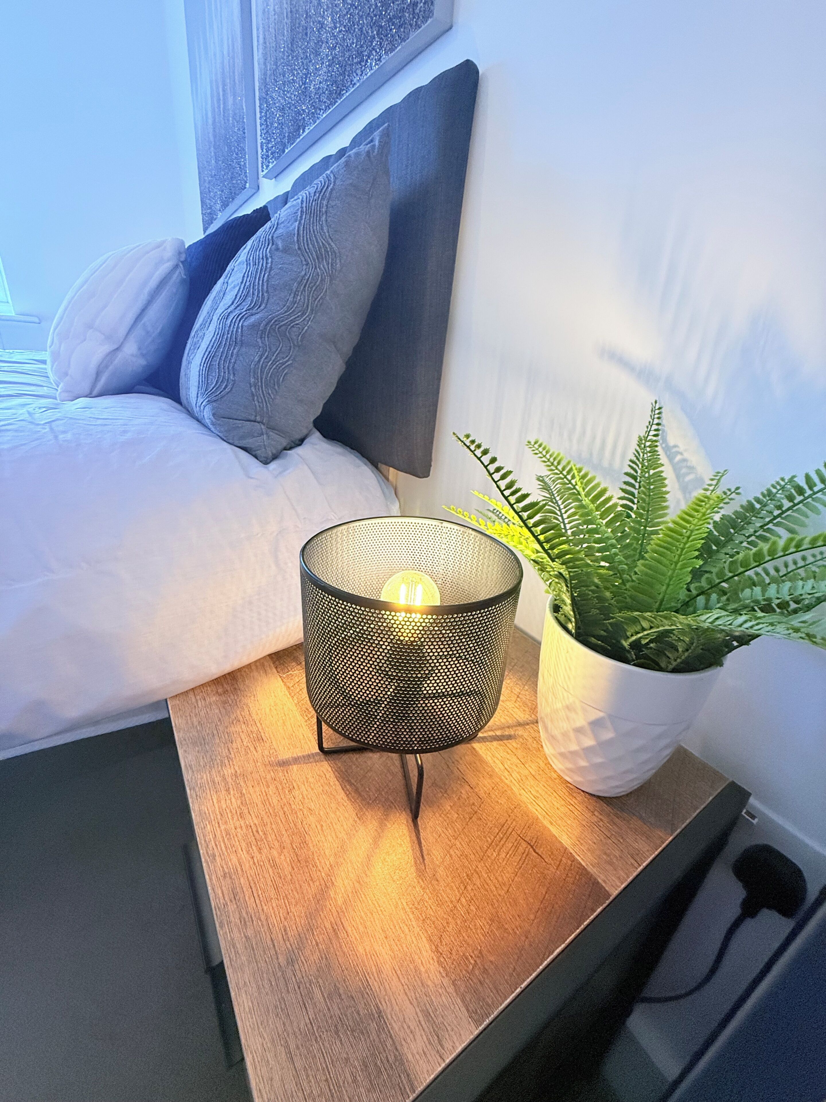 En seng med sengegavl, et natbord med en metal lampe, og en pottefern.