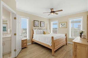 Quarto com cama de madeira, ventoinha no teto e duas janelas com persianas.