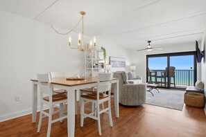 A dining area with a wooden table and white chairs, a chandelier, a bookshelf, and a view of the ocean through a sliding glass door. - Ocean Breeze West 705 (Pensacola)