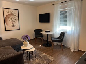 Ein modernes Wohnzimmer mit einer Couch, einem kleinen runden Tisch, zwei Stühlen, einem Flachbildfernseher und einem Fenster mit durchsichtigen Vorhängen.