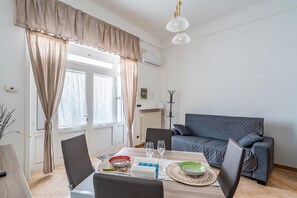 A dining area with a table set for two, a sofa, and a window with curtains. - Casa Alessia by Rentbeat (Cisternino)