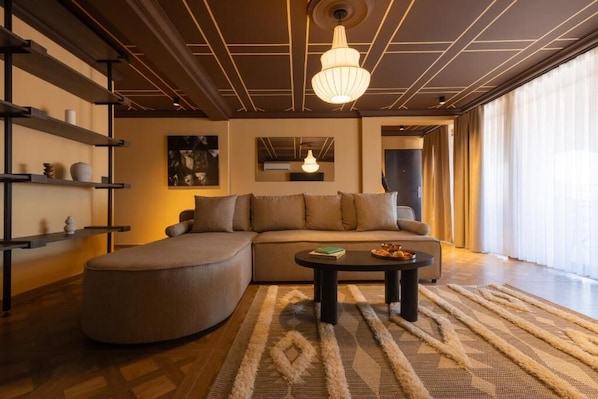 Una sala de estar moderna con un sofá modular, un reposapiés redondo, una mesa de centro y un amplio ventanal con cortinas.