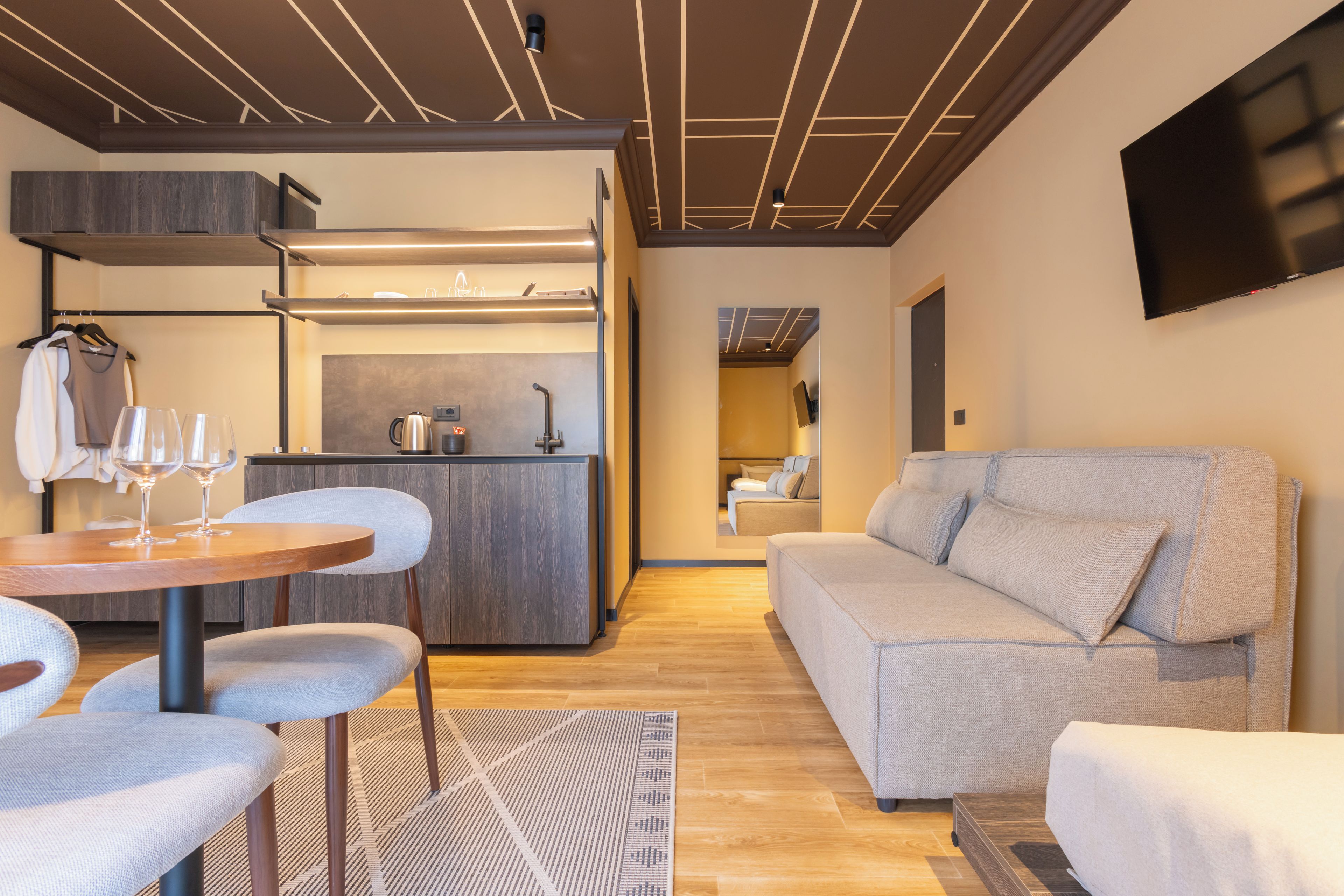 A modern interior with a sofa, dining table, and chairs, a kitchen area, and a flat-screen TV mounted on the wall.