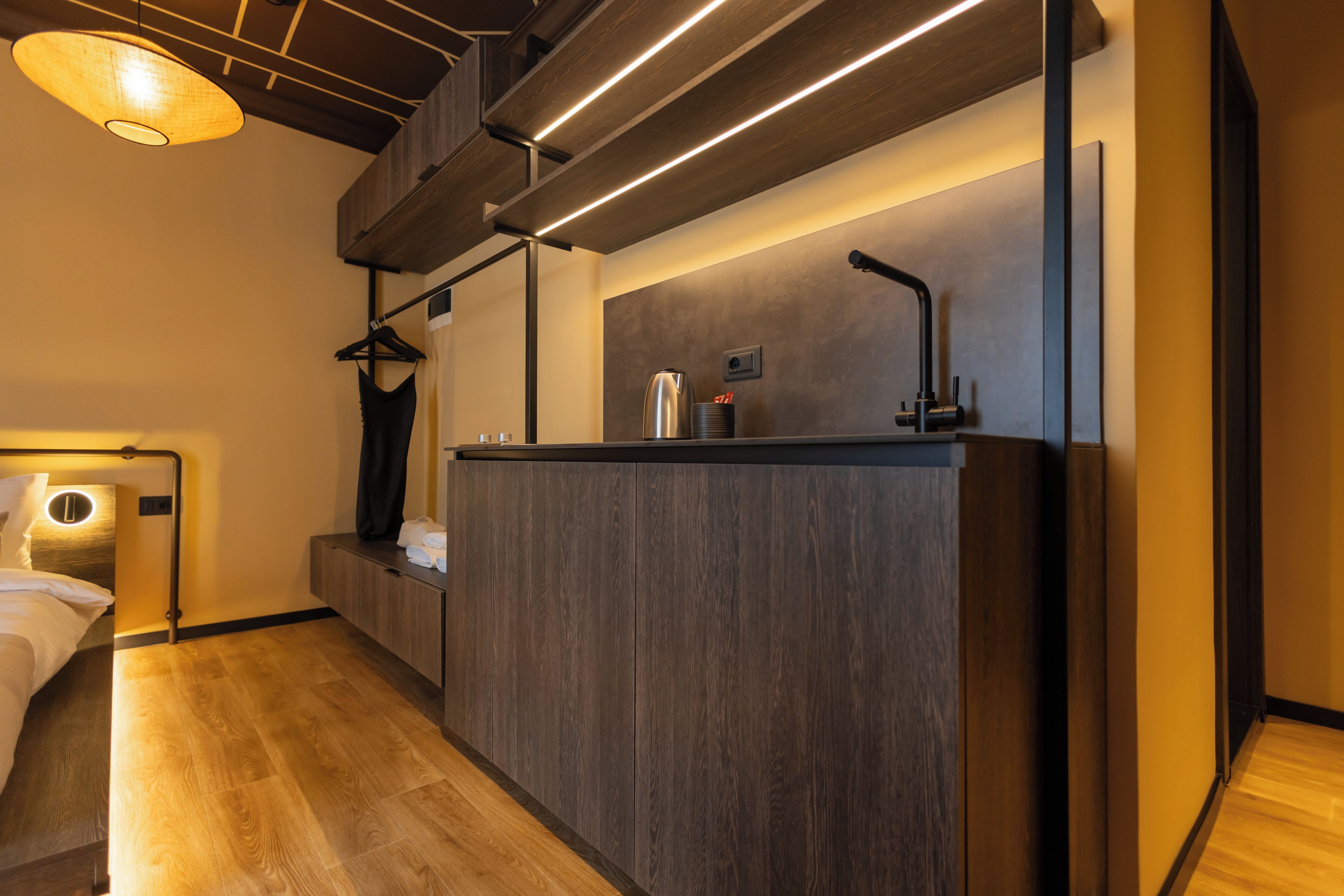 A modern hotel room with a wooden floor, a bed with a circular light, a dark wooden desk, and a hanging coat rack.