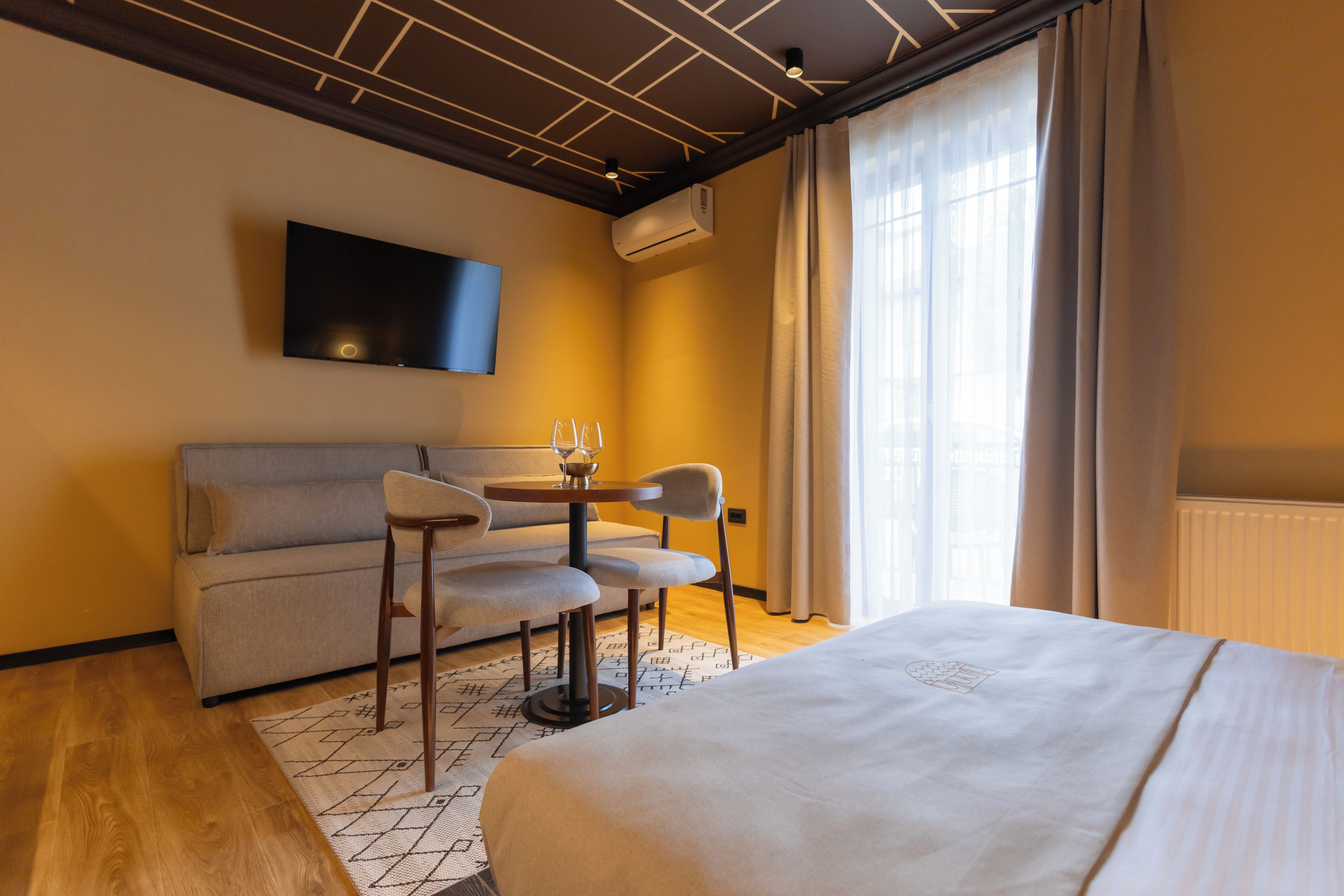 Habitación de hotel con cama, sofá, una pequeña mesa con sillas, televisor montado en la pared y ventana con cortinas.