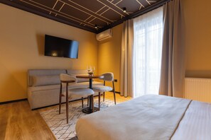 Habitación de hotel con cama, sofá, una pequeña mesa con sillas, televisor montado en la pared y ventana con cortinas.