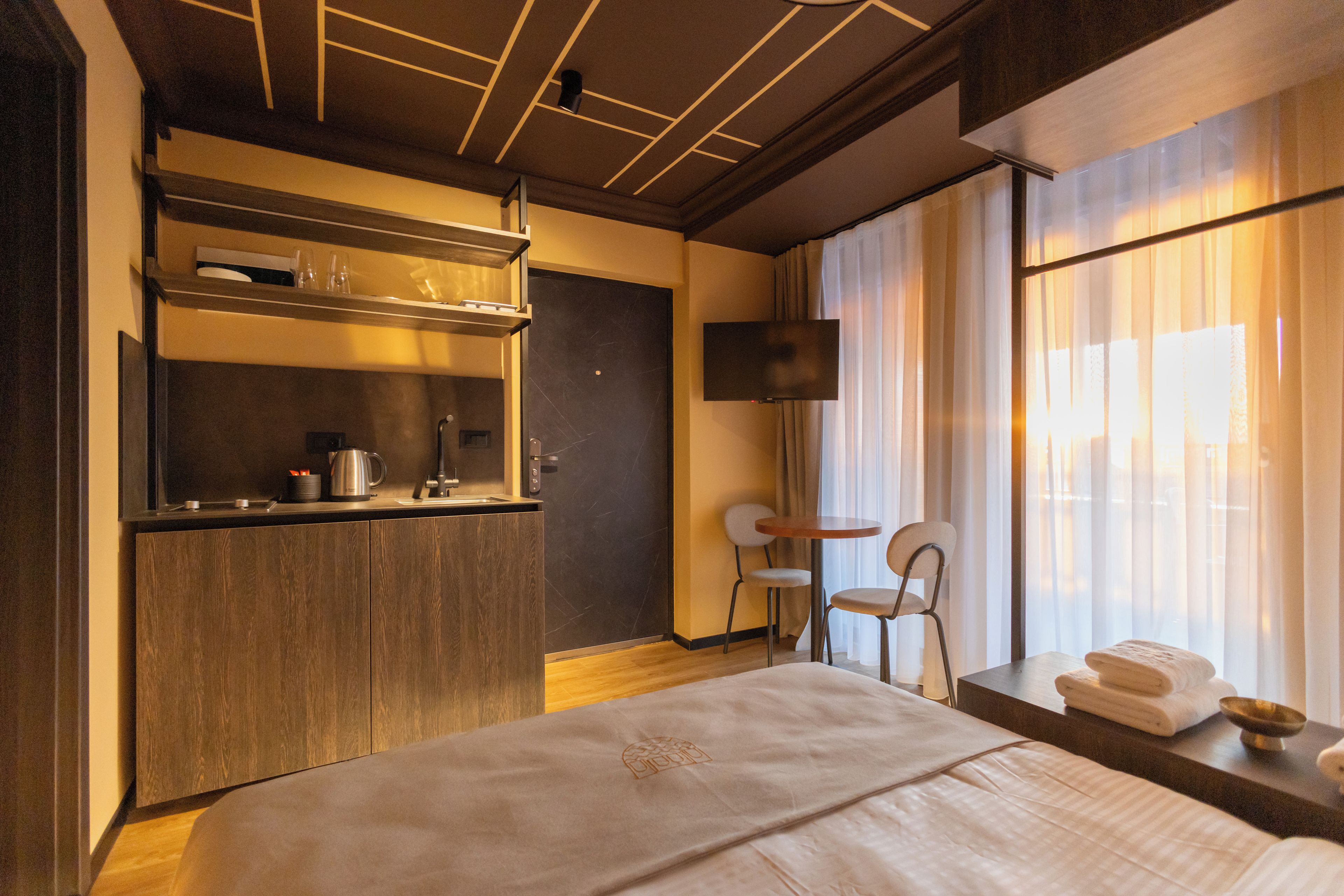 A hotel room with a bed, a small kitchenette, a TV, and a window with curtains.