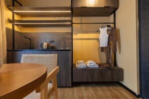 Una habitación compacta con una mesa de madera, una silla, un banco empotrado con toallas, una percha para ropa y una kitchenette.