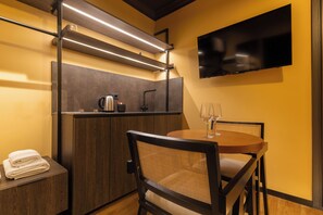 Habitación de hotel compacta con kitchenette, un televisor de pantalla plana en la pared, una mesa de comedor con sillas y una toalla en la mesita de noche.