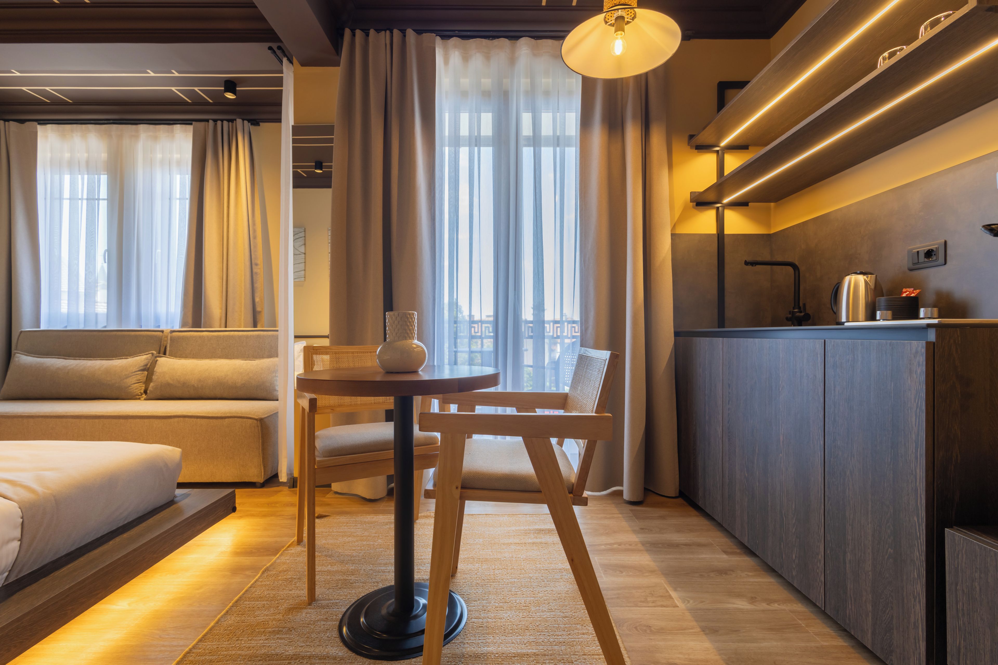 A modern hotel room with a sofa, a small round table, a chair, a kitchen area with a countertop and cabinets, and large windows with curtains.
