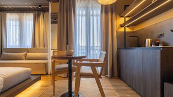 A modern hotel room with a sofa, a small round table, a chair, a kitchen area with a countertop and cabinets, and large windows with curtains.