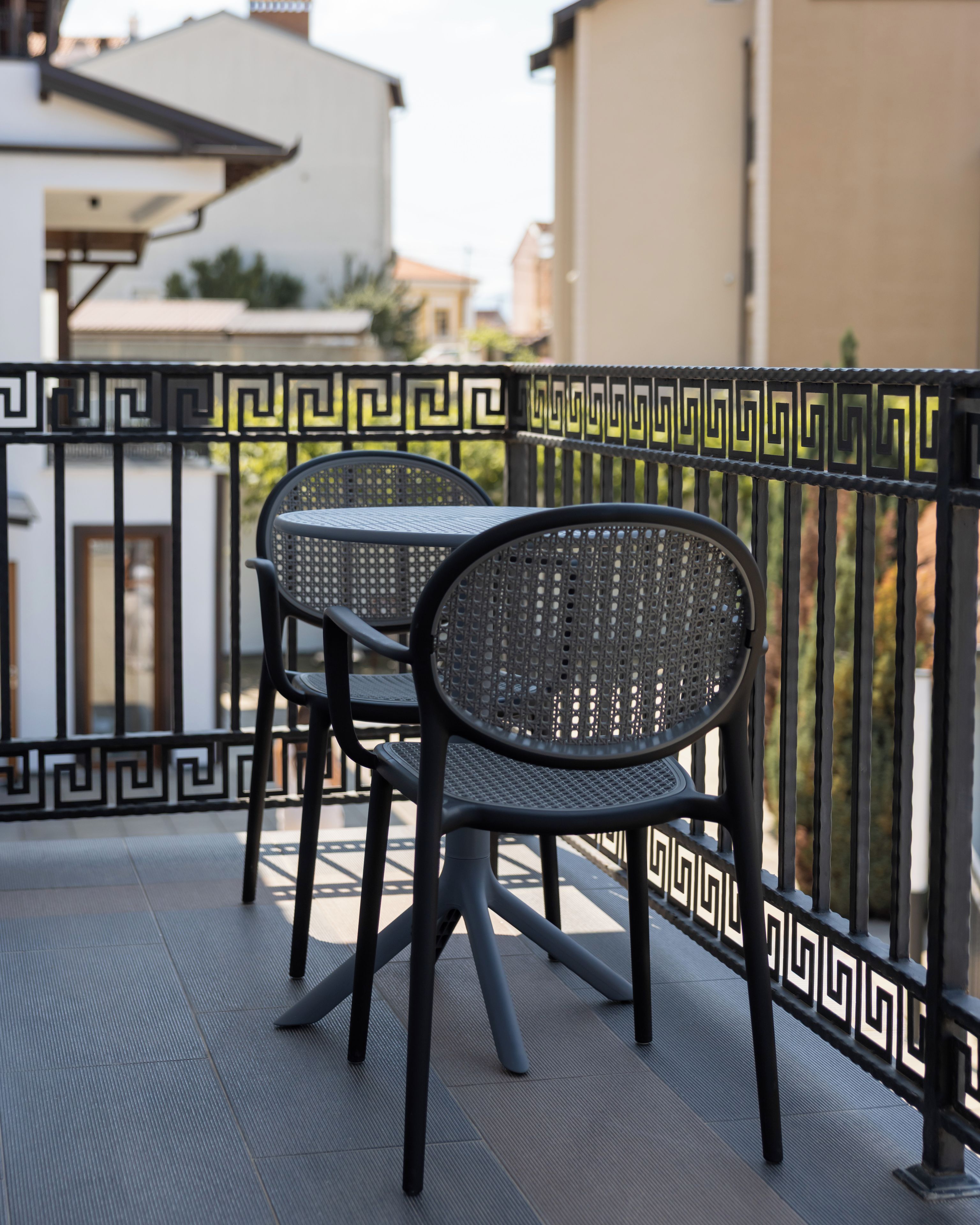 A balcony with a black metal table and two chairs, a decorative black and white railing, and a building in the background.