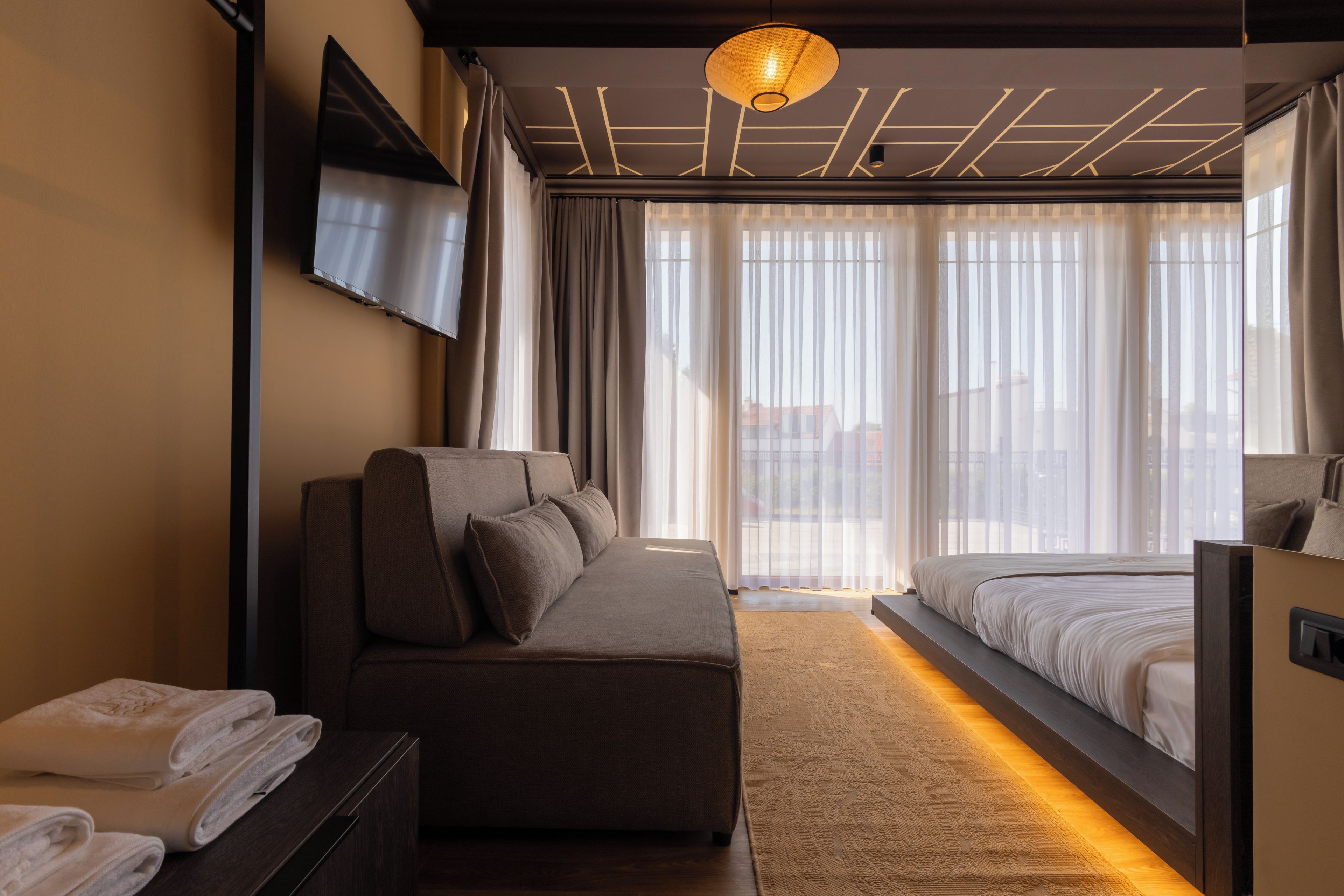 A modern hotel room with a sofa, a bed, a flat-screen TV, and a large window with sheer curtains.