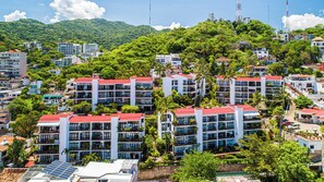 A hillside residential area with multi-story buildings, some with red roofs, surrounded by lush greenery and a mountain in the background. - SPECTACULAR views of Banderas Bay One Bedroom Condo in Centro (Puerto Vallarta)