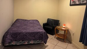 Ein kleiner, ordentlicher Raum mit einem Bett, einem Sessel, einem Tisch mit einem Heizgerät und einem Bild an der Wand.