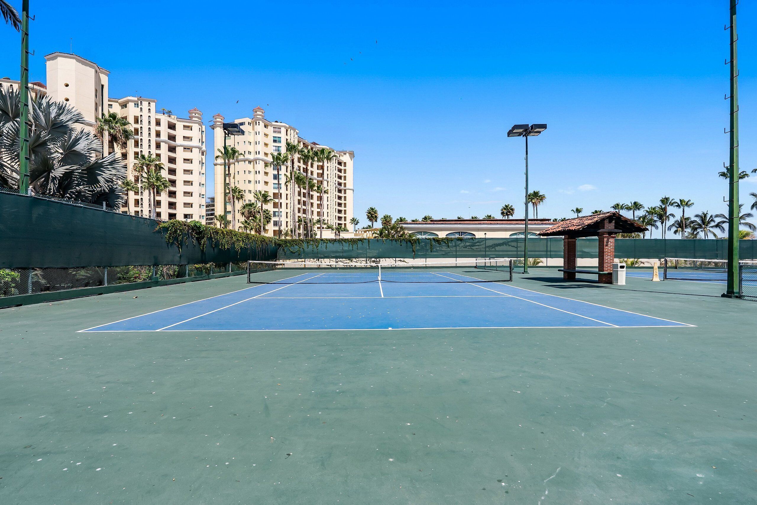 A tennis court with a net, surrounded by a fence, palm trees, and a building in the background.