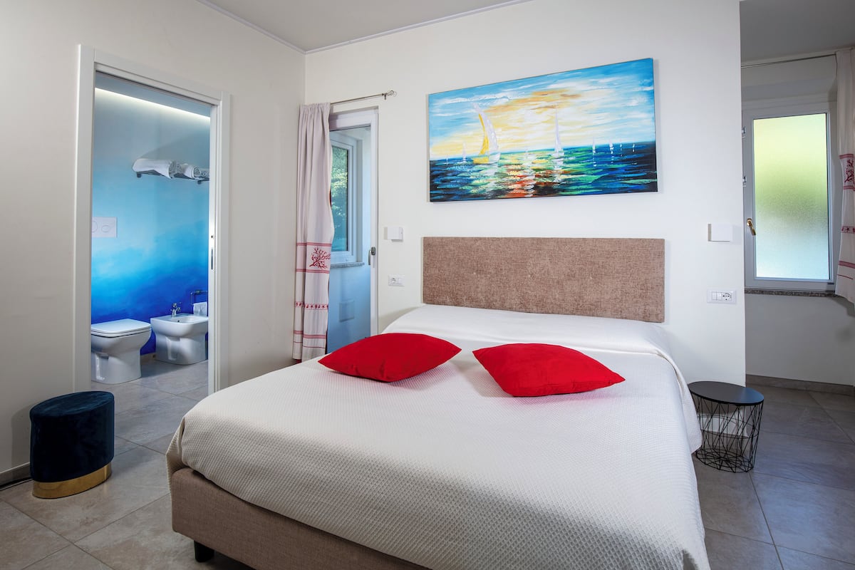 A hotel room with a bed, a painting of a sailboat, a bathroom with a blue accent wall, and a small stool.