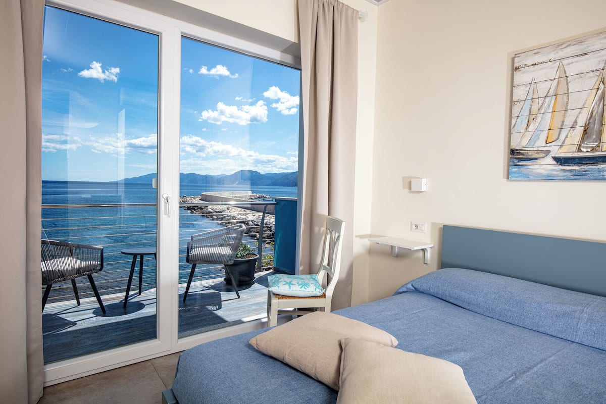A room with a bed, a chair, a table, and a view of the sea through a glass door.