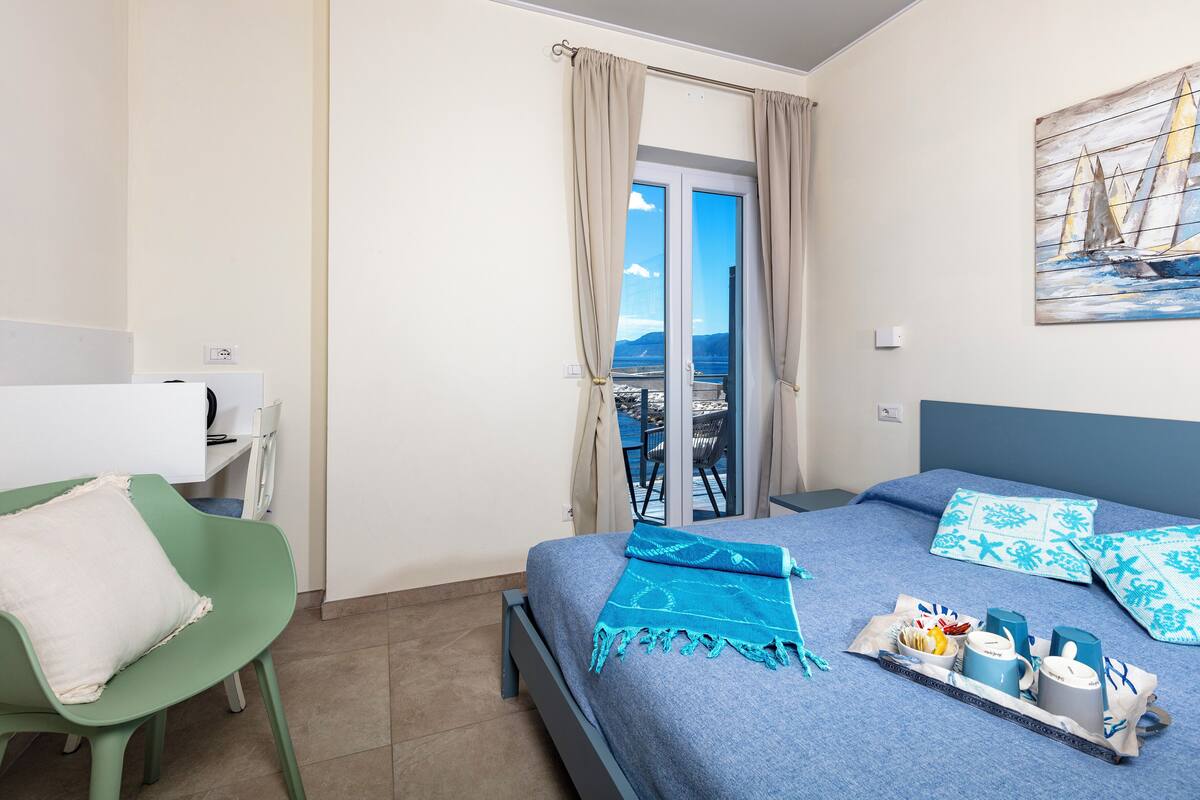 A bedroom with a bed, a green chair, a table with a tray of items, and a balcony with a view.