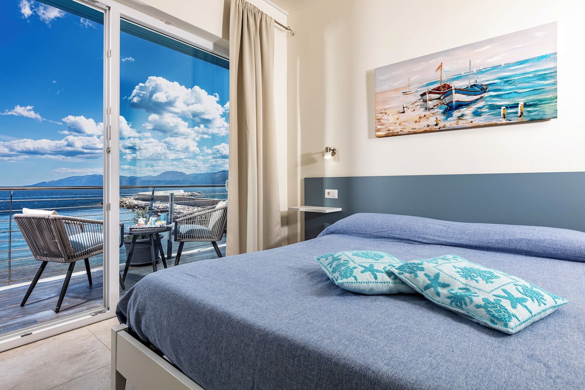 A balcony with a view of the sea, a bed with blue bedding, and a painting of a boat on the wall.
