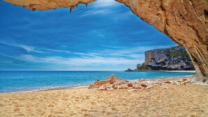Una playa con una formación rocosa natural en forma de arco sobre la arena, cielo azul despejado y mar en calma.