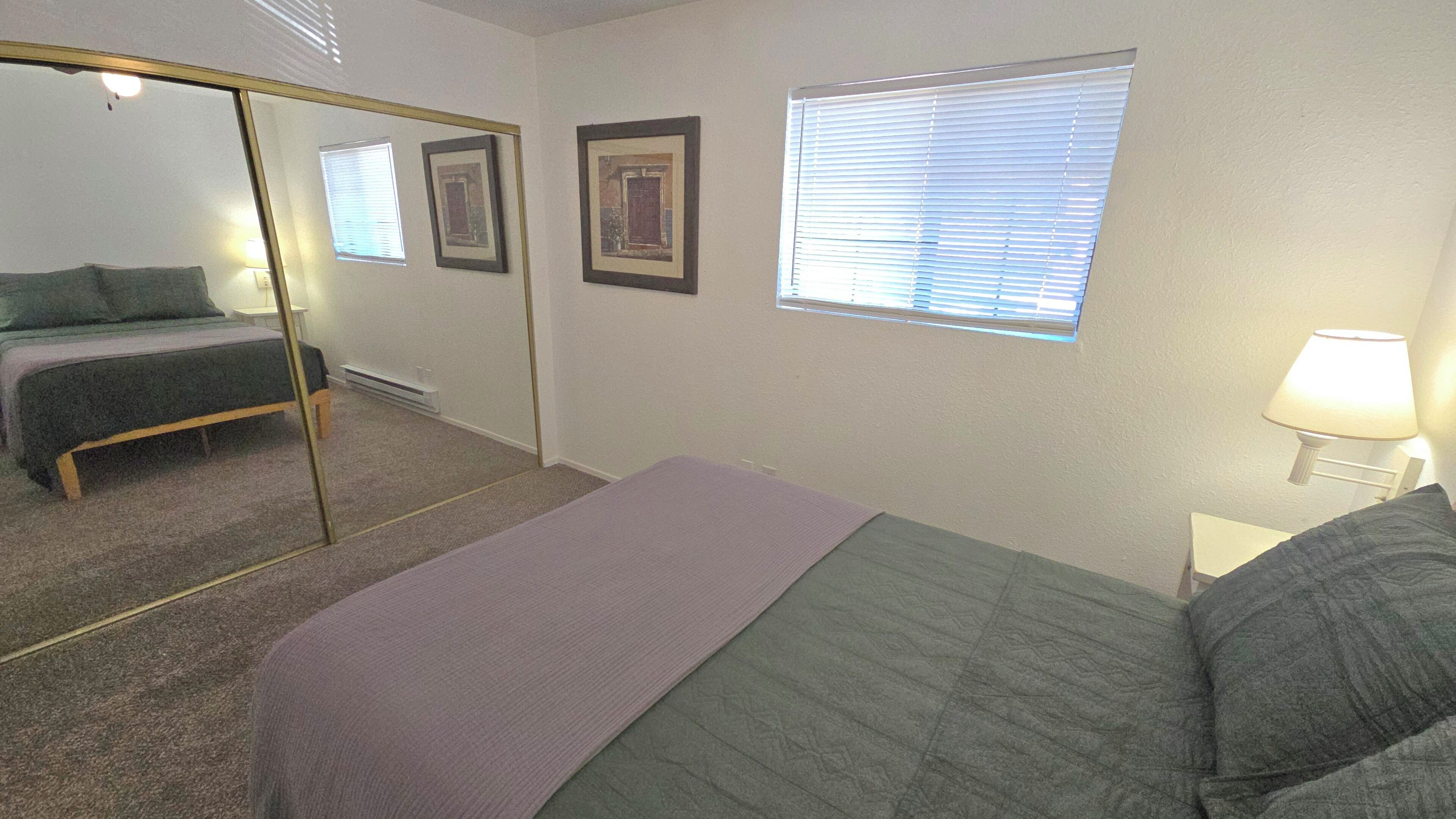 Une chambre à coucher comprenant un lit, un miroir, deux tableaux encadrés, une fenêtre avec des stores et une table de chevet avec une lampe.