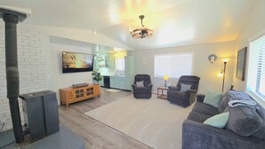 A living room with a fireplace, a sofa, two armchairs, a TV, and a kitchen in the background. - Large Home, Sleeps 13, Hot Tub, Starlink, Air Conditioning (Eagar)