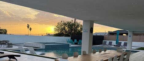 Un espace aménagé au bord de la piscine, avec une table en bois et des chaises blanches, un coin salon abrité et une vue sur le coucher du soleil.