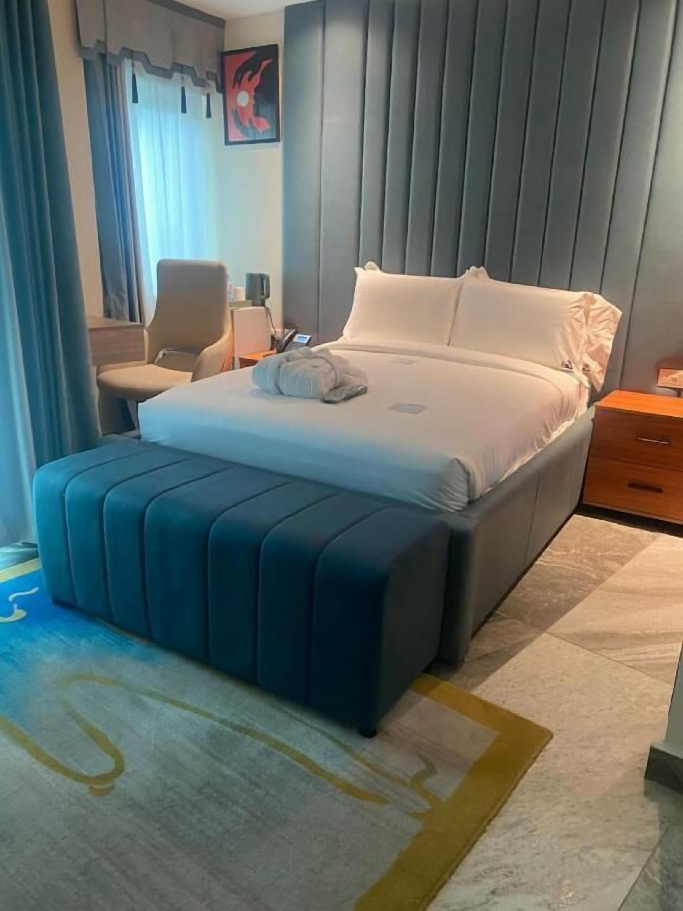 A hotel room with a bed, a chair, a bedside table, and a wall with a painting.
