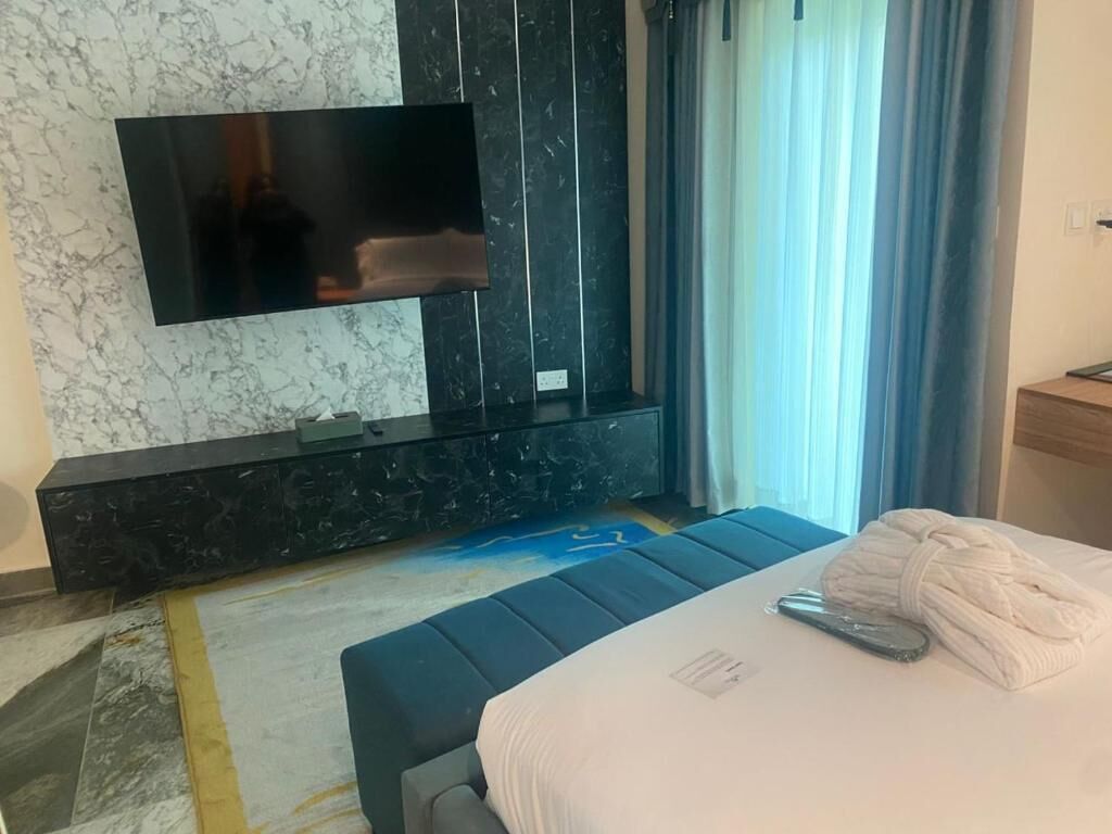 A hotel room with a flat-screen TV mounted on a dark marble wall, a bed with a white comforter, and a small wooden side table.