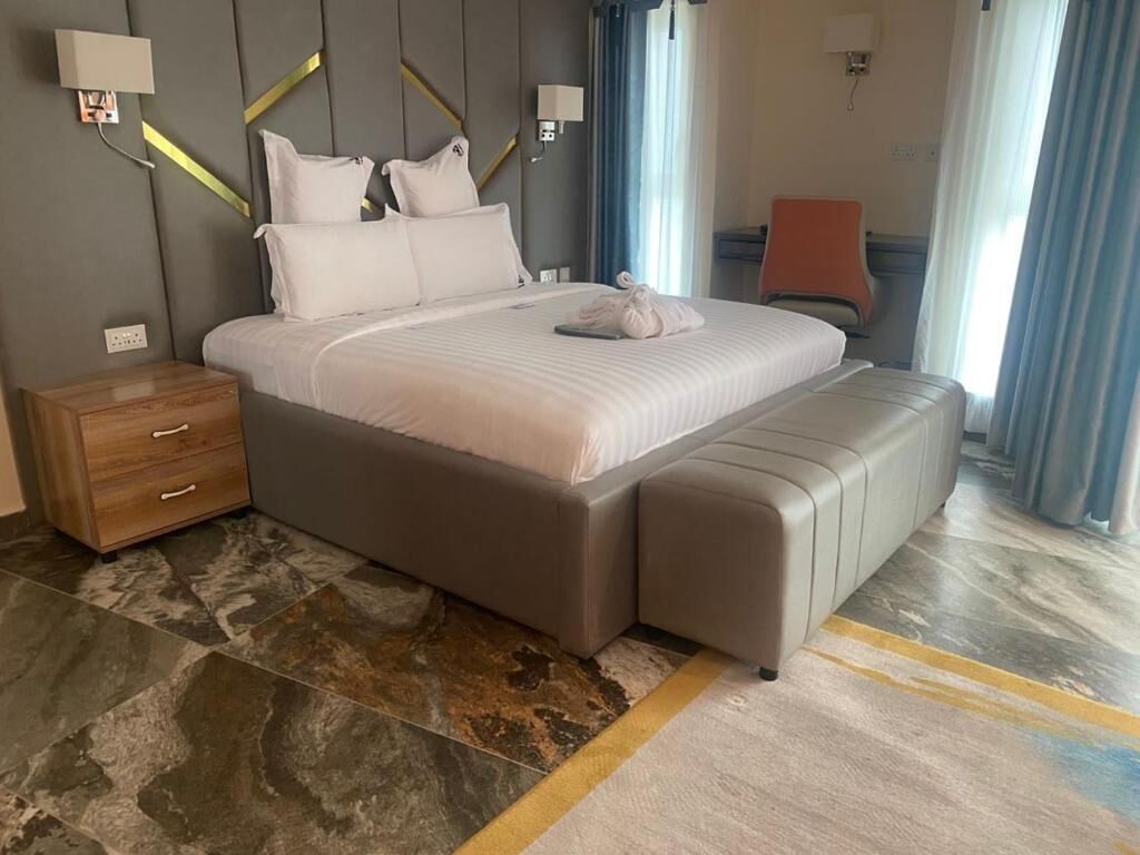 Une chambre d’hôtel moderne avec un grand lit, une table de chevet en bois, un banc et une vue sur la ville grâce à de grandes fenêtres.