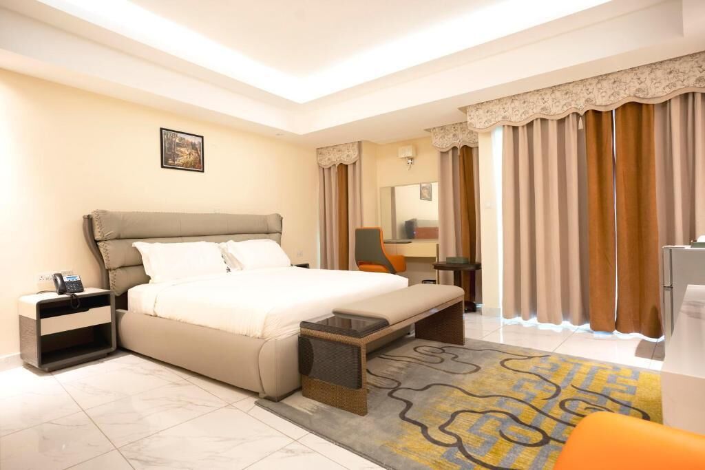 A hotel room with a bed, bedside table, lamp, and a view of the outside through curtains.