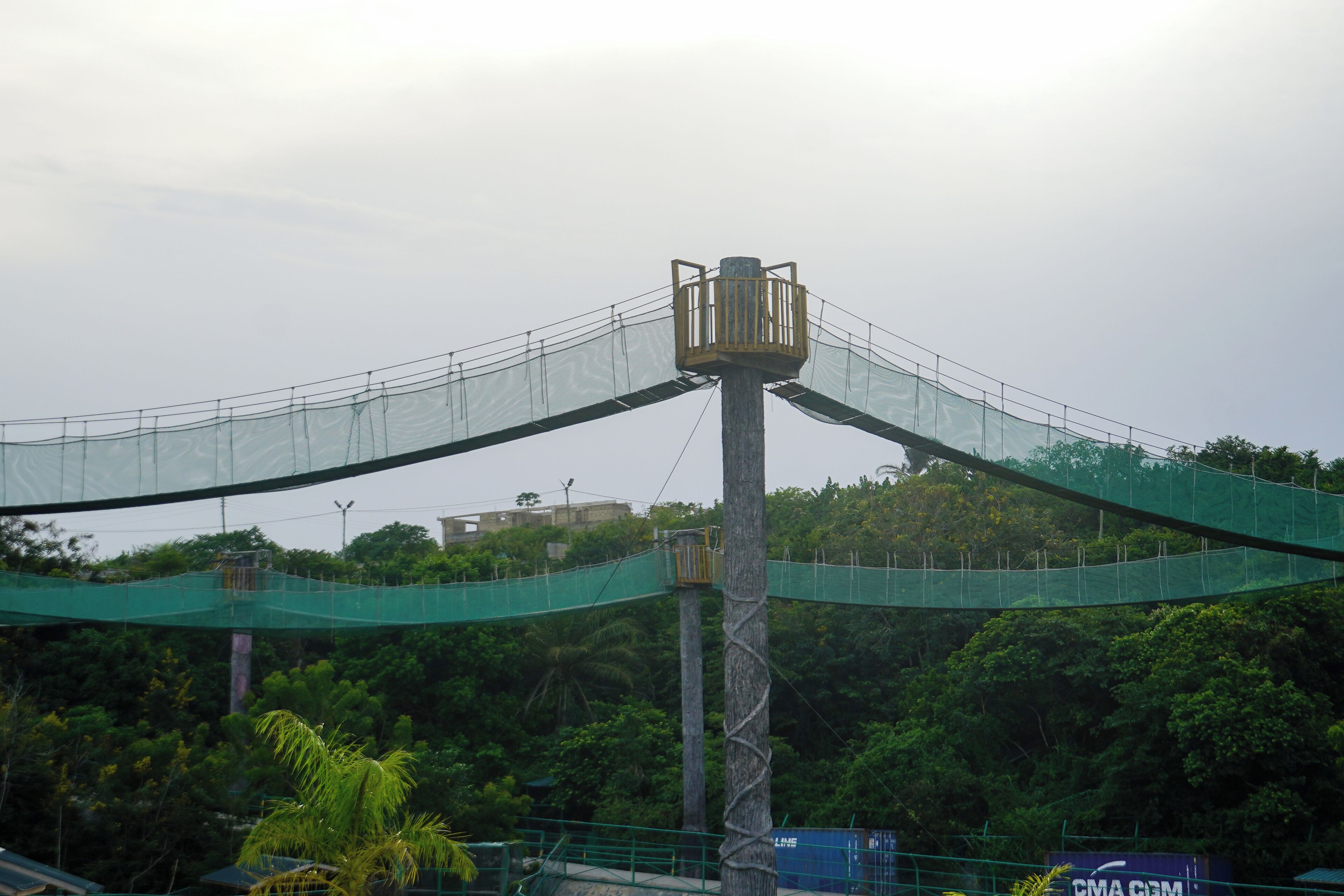 A suspension bridge with a central observation tower, surrounded by dense greenery and a distant building.