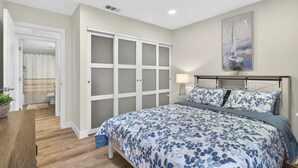 Une chambre à coucher avec un lit, une armoire, un tableau et une salle de bain visible à travers une porte ouverte.