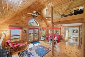 A wooden cabin interior with a loft bedroom, a dining area, and a view of the outdoors.