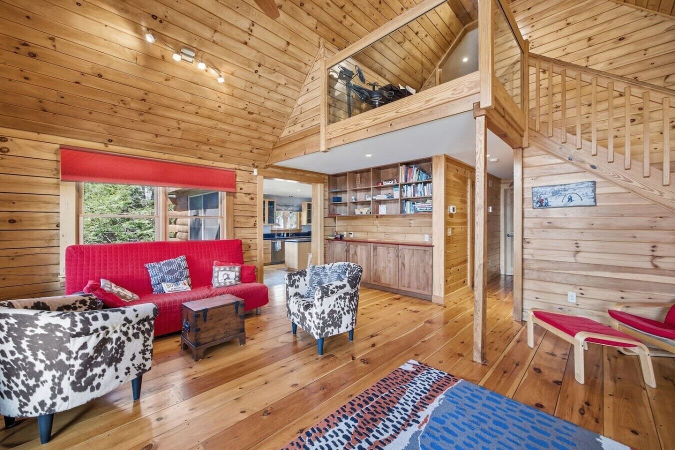 A wooden cabin interior with a red sofa, patterned chairs, a wooden staircase, and a kitchen area.