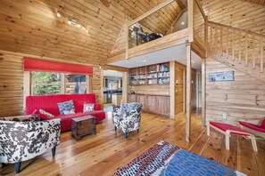 A wooden cabin interior with a red sofa, patterned chairs, a wooden staircase, and a kitchen area.