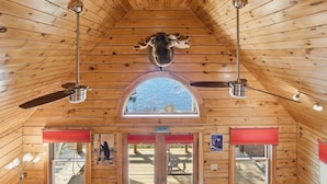 Ein Holzblockhaus-Interieur mit schrägem Dach, zwei Deckenventilatoren, einem großen Bogenfenster und einem an der Decke befestigten Bärenkopf.
