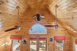 A wooden cabin interior with a sloped ceiling, two ceiling fans, a large arched window, and a bear head mounted on the ceiling.
