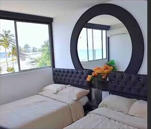 A hotel room with two beds, a large mirror, a window with a view of palm trees and the sea, and a vase with flowers. - CRESPO OCEAN VIEW APT (Cartagena de Indias)