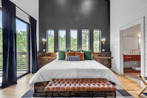 Ein großes Bett mit einer Bank, eine Holzbank, ein großes Fenster und Blick auf Bäume.