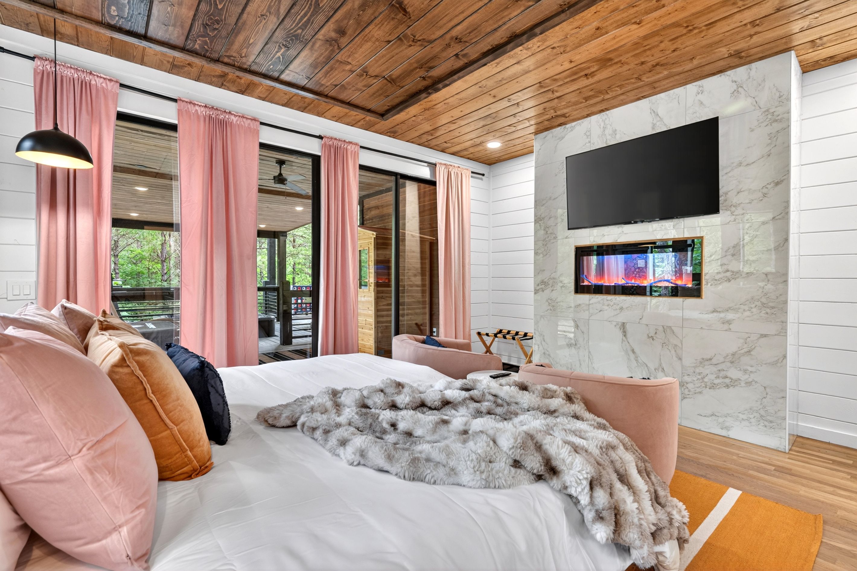 A modern bedroom with a large bed, pink curtains, a TV mounted on a marble wall, and a wooden ceiling.